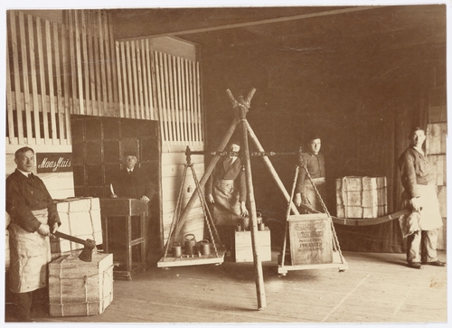 Gemeentelijk Handelsentrepôt pand 'Zondag', interieur, adres: Cruquiusweg 25. datering zo rond 1905. Links staat een man met een bijl bij de kist, daarachter een man achter een lessenaar, iemand bij een grote weegschaal en twee mannen die een kist tillen. Collectie Beeldbank SAA.   